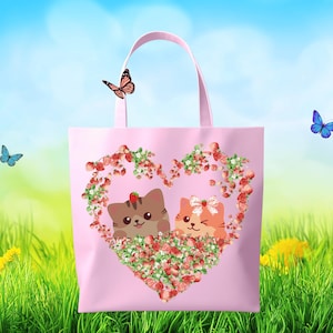 May include: A pink tote bag featuring two cartoon cats inside a heart-shaped floral design. The bag has a light pink handle and is set against a backdrop of green grass, blue sky, and butterflies.