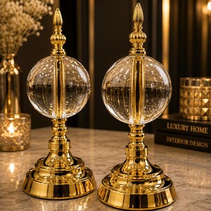 Op de afbeelding: Twee decoratieve tafelornamenten met een heldere glazen bol op een gepolijste goudkleurige metalen voet. Elk stuk heeft een verticale gouden staaf die door het midden van de bol loopt, bekroond met een puntige, geribbelde gouden knop.