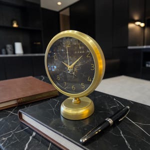Puede incluir: Un reloj de mesa analógico redondo con marco y base de metal dorado cepillado. La esfera tiene un fondo negro con vetas blancas similares al mármol y números dorados finos del 1 al 12. Tiene dos manecillas doradas que indican la hora.