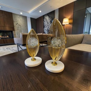 Op de afbeelding: Twee decoratieve sculpturen met goudkleurige metalen draadframes in de vorm van langwerpige bladeren. Elke sculptuur bevat een heldere, ronde glazen bol met interne luchtbellen, gemonteerd op een ronde voet van wit marmer.