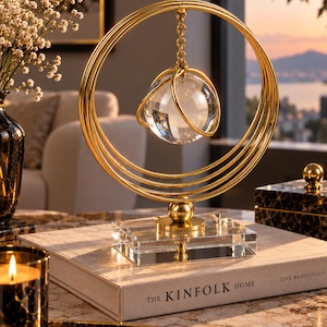 Op de afbeelding: Een decoratief sculptuur van goudkleurig metaal met een reeks concentrische ringen op een rechthoekige voet van helder glas. Een glazen bol hangt aan een gouden ketting aan de ringen en is omsloten door een kleinere gouden ring.
