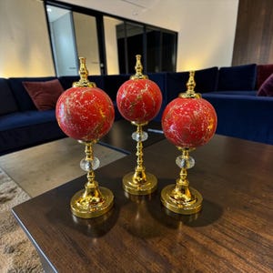 Op de afbeelding: Drie decoratieve ornamenten met bolvormige rode bovenkanten met gouden marmerpatronen. Elk stuk staat op een goudkleurige metalen voet met een heldere gefacetteerde kristallen kraal op de centrale steel onder de bol.