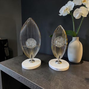 Op de afbeelding: Twee decoratieve sculpturen met zilverkleurige metalen draadframes in de vorm van langwerpige bladeren. Elke sculptuur bevat een heldere glazen bol met interne luchtbellen, gemonteerd op een solide ovale voet van wit marmer.