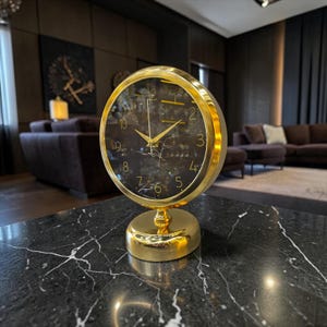 Puede incluir: Un reloj de mesa redondo con marco y base de oro pulido. La esfera es negra con un sutil patrón de vetas blancas similar al mármol, con números dorados del 1 al 12 y manecillas doradas a juego para las horas, los minutos y los segundos.