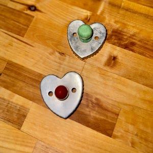 May include: Two heart-shaped cookie cutters with a metal finish. One cutter has a red center, and the other has a green center. The cutters are on a wooden surface.