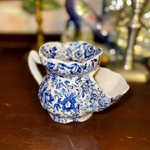 May include: A ceramic shaving mug featuring a blue and white floral pattern. The mug has a scalloped rim, a side handle, and a built-in soap compartment on the front. The exterior is decorated with intricate botanical illustrations in blue ink.