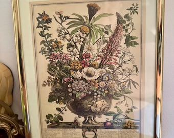 Puede incluir: Una impresión botánica enmarcada con una ilustración detallada de diversas flores y follaje dispuestas en una urna decorativa. La impresión tiene un fondo crema y está encerrada en un marco de metal dorado. La palabra "DICIEMBRE" es visible en la parte inferior.