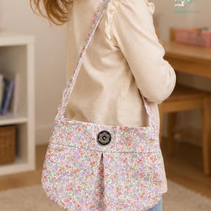 May include: A shoulder bag featuring a small floral pattern in shades of pink, purple, and green. The bag has a single long strap and a central black button closure. The fabric displays a pleated design on the front panel of the bag.