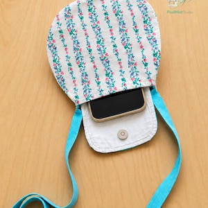 May include: A white fabric crossbody bag featuring a vertical floral pattern in pink, blue, and green. The bag has a bright turquoise strap and a magnetic snap closure. A black smartphone is tucked inside the open pouch. Text reads PixelMintStudio and STEP BY STEP GUIDE BEGINNER FRIENDLY.