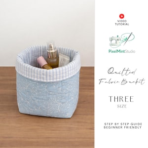Puede incluir: Un cubo de tela acolchada azul claro con un estampado floral blanco y un borde a rayas. El cubo contiene varios productos cosméticos. El texto en el lateral dice "Quilted Fabric Bucket" y "THREE SIZE".
