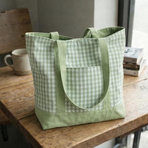 May include: A green and white gingham tote bag with a solid green base, pocket, and straps. The bag is made of fabric and has a rectangular shape. The tote bag is sitting on a wooden table.