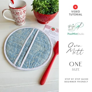 May include: A round, light blue oven mitt with a white floral pattern and white trim. Two white stripes cross the mitt. The words "Oven Mitt" and "ONE SIZE" are visible. A red spatula and a mug are also in the image.