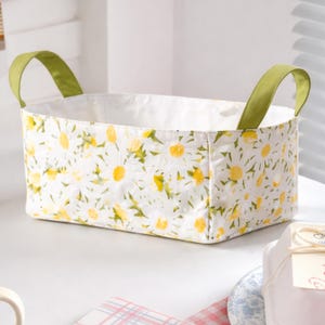 May include: A rectangular fabric storage basket with a white background and a daisy floral pattern. The basket has green handles and a white interior. The basket is sitting on a white surface.