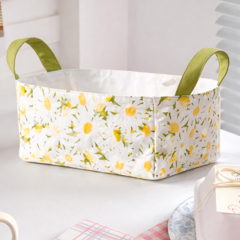 May include: A rectangular fabric storage basket with a white background and a daisy floral pattern. The basket has green handles and a white interior. The basket is sitting on a white surface.