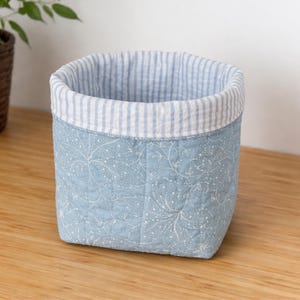 May include: A square, quilted fabric basket with a light blue floral pattern on the exterior. The top edge features a striped blue and white fabric. The basket is sitting on a wooden surface.