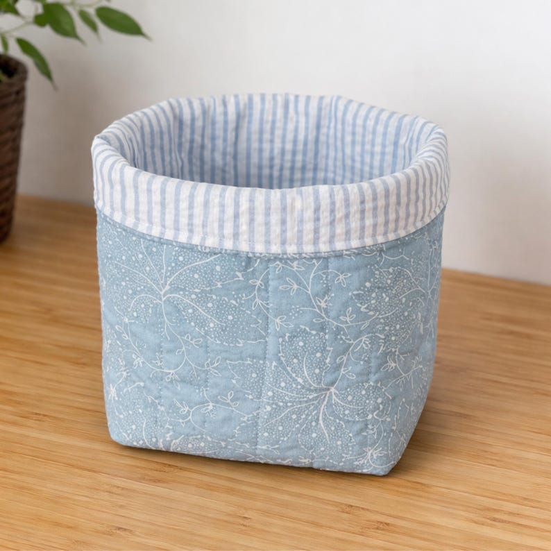 May include: A square, quilted fabric basket with a light blue floral pattern on the exterior. The top edge features a striped blue and white fabric. The basket is sitting on a wooden surface.