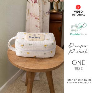 Könnte beinhalten: Eine weiße Windeltasche mit Reißverschluss und Griff, verziert mit kleinen gelben und grünen Mustern. Die Tasche hat ein Etikett mit der Aufschrift "AkkoBaby". Sie steht auf einem kleinen Holztisch.