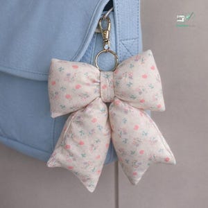May include: A cream-coloured, floral bow-shaped keyring with a gold-coloured clasp is attached to a light blue bag. The bow features a delicate pattern of pink and purple flowers.