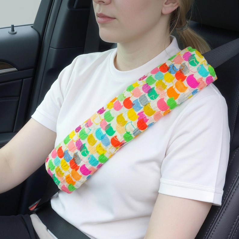 Puede incluir: Una funda colorida para cintur&oacute;n de seguridad con un patr&oacute;n de c&iacute;rculos multicolores. La funda est&aacute; envuelta alrededor de un cintur&oacute;n de seguridad y descansa sobre el hombro de una persona. El fondo muestra el interior de un coche.