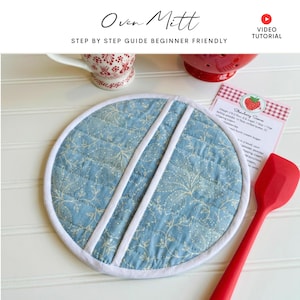 May include: A round, quilted oven mitt with a light blue floral pattern and white trim. Two white stripes cross the mitt diagonally. The text "Oven Mitt" and "Step by Step Guide Beginner Friendly" are visible.
