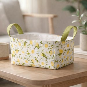 May include: Rectangular fabric storage basket with a white daisy floral pattern and green handles. The basket is on a wooden table. The interior is white. The handles are a light green color.