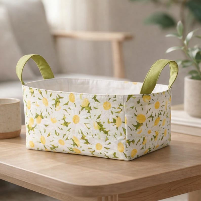 May include: Rectangular fabric storage basket with a white daisy floral pattern and green handles. The basket is on a wooden table. The interior is white. The handles are a light green color.
