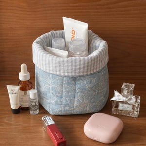 May include: A blue and white floral patterned fabric basket with a striped rim, filled with cosmetic products. Several beauty products, including a perfume bottle, lipstick, and skincare items, are arranged around the basket on a wooden surface.