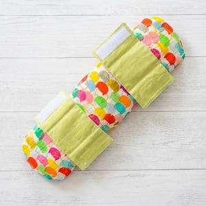 Puede incluir: Una envoltura para beb&eacute; colorida con un patr&oacute;n de formas multicolores. La envoltura tiene tres correas verde claro con cierres de velcro blancos. Est&aacute; sobre una superficie de madera blanca.