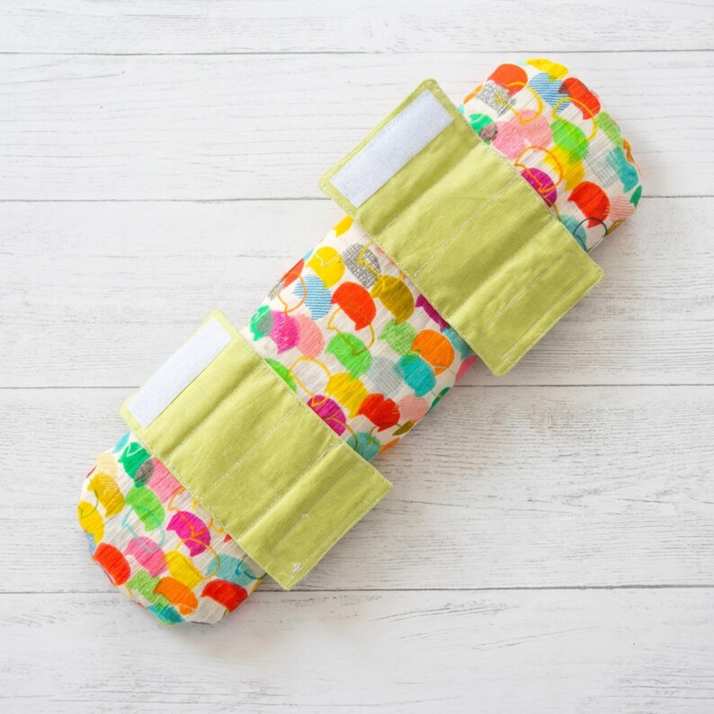 Puede incluir: Una envoltura para beb&eacute; colorida con un patr&oacute;n de formas multicolores. La envoltura tiene tres correas verde claro con cierres de velcro blancos. Est&aacute; sobre una superficie de madera blanca.