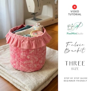 Puede incluir: Un cubo de tela rosa con la parte superior con volantes, lleno de rollos y retales de tela. El cubo tiene un estampado floral y está sobre una superficie beige. El texto "Fabric Bucket" y "THREE SIZE" son visibles.