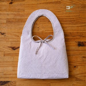 May include: A light purple and white gingham fabric bib featuring a rounded neckline with a matching fabric tie closure at the center. The bib has a simple, flat construction with a clean hem along the bottom edge. Text at the bottom reads STEP BY STEP GUIDE BEGINNER FRIENDLY.