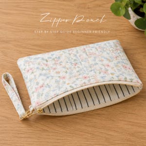 May include: A floral print zipper pouch with a gold zipper and wrist strap. The pouch features a quilted design with a white background and pink and blue flowers. The interior lining has a striped pattern. Text on the image reads "Zipper Pouch" and "Step by Step Guide Beginner Friendly."