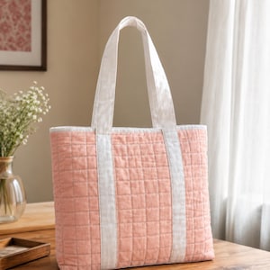 May include: A quilted tote bag in a soft peach color featuring a grid pattern and two white fabric handles. The bag has a white trim along the top edge. The text "STEP BY STEP GUIDE BEGINNER FRIENDLY" is printed in white at the bottom of the image.