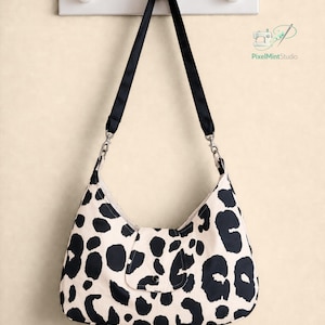 May include: A hobo-style shoulder bag featuring a cream-coloured base with a black leopard print pattern. The bag includes a black fabric strap attached with silver-toned metal clips and a central flap closure with a single black snap button.