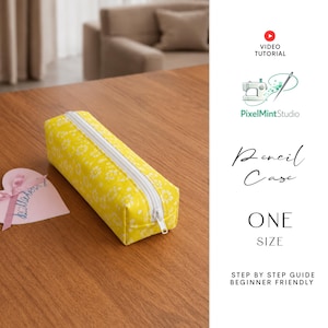 May include: A yellow pencil case with a white floral pattern and a white zipper. The pencil case is rectangular and sits on a wooden surface. A small heart-shaped card with a pink ribbon is to the left of the pencil case. The text "Pencil Case" is visible.