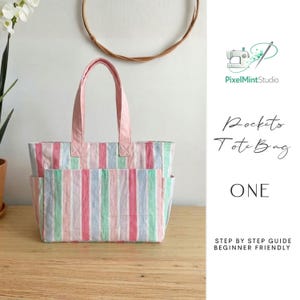 May include: A pastel striped tote bag with pink handles and a front pocket. The bag features vertical stripes in pink, blue, and green. The text "Pocket Tote Bag" and "ONE" are visible on the right side of the image.