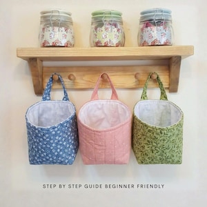 Può includere: Tre cestini sospesi in tessuto a motivi floreali blu, pois rosa e foglie verdi. I cestini sono appesi sotto una mensola di legno con tre barattoli di vetro con carta floreale all'interno. Il testo "STEP BY STEP GUIDE BEGINNER FRIENDLY" è in basso.