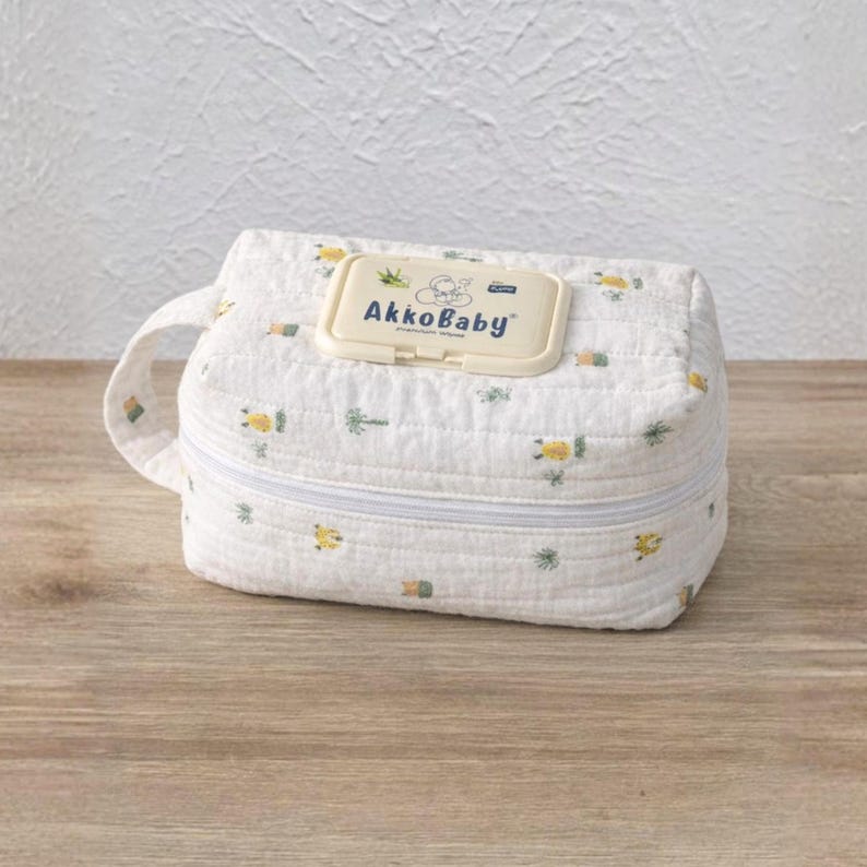 May include: White fabric baby wipes container with a zipper closure and a handle. The container has a pattern of yellow and green floral designs. The lid of the wipes container has the text "AkkoBaby".