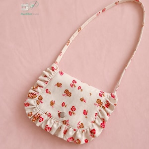 Op de afbeelding: Een kleine schoudertas van gebroken witte stof met een herhalend bloemenpatroon in rood- en bruintinten. De tas heeft een afgeronde klep met een geplooide rand, een centrale drukknoopsluiting en een bijpassende schouderband van dezelfde stof.