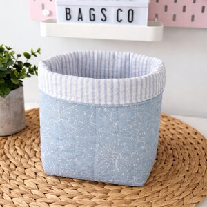 May include: A square, light blue fabric basket with a white floral pattern and a striped interior. The basket sits on a woven mat, with the text "BAGS CO" visible in the background.
