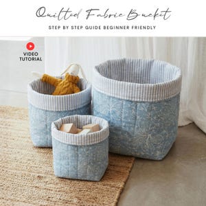 Può includere: Tre secchi in tessuto trapuntato di diverse dimensioni. I secchi sono azzurri con un motivo floreale bianco e una fodera a righe. Il testo "Quilted Fabric Bucket" e "Step by Step Guide Beginner Friendly" è in alto.