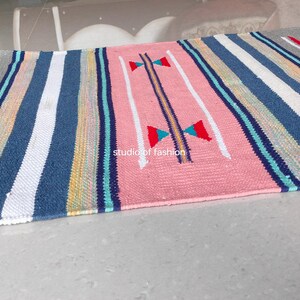Può includere: Un tappeto rettangolare tessuto con un motivo a righe nei toni del grigio, blu, bianco, rosa e accenti di rosso, verde e giallo. Il tappeto ha un bordo sfrangiato e il testo "studiof fashion" è visibile.
