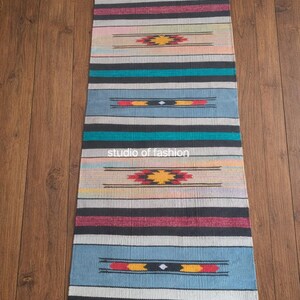 May include: A rectangular, woven runner rug with a colorful striped pattern. The rug features bands of blue, black, red, and tan, with geometric designs in the center. The text "studio of fashion" is printed on the rug.