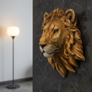 Op de afbeelding: Een driedimensionaal wandsculptuur van een leeuwenkop met een goudbruine afwerking. Het object is voorzien van gedetailleerde vachttextuur, een prominente manen en realistische gelaatstrekken, gemonteerd op een donker, marmerachtig oppervlak.