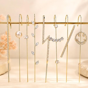 May include: A collection of eight gold-tone ear crawlers and climbers featuring various designs including a pearl, clear crystals in linear, leaf, lightning bolt, and smiley face shapes, all suspended from a thin horizontal gold-tone metal bar.