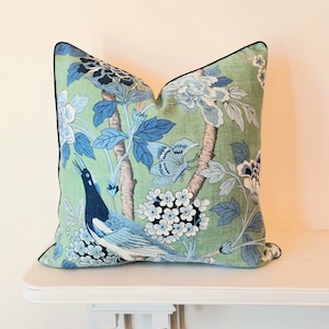 Könnte beinhalten: Ein quadratisches Zierkissen mit hellgrünem Hintergrund und einem blau-weißen botanischen Druck. Das Muster zeigt einen Vogel, einen Schmetterling und verschiedene florale Elemente. Das Kissen ist mit einer schmalen schwarzen Paspelierung versehen.