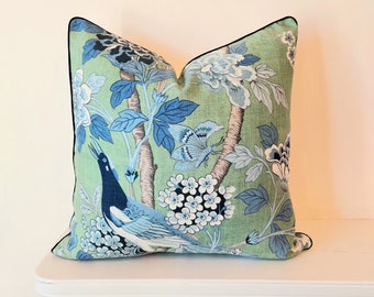 May include: A square throw pillow featuring a light green background with a blue and white botanical print. The pattern includes a bird, a butterfly, and various floral elements. The pillow is finished with a thin black piped edge trim.