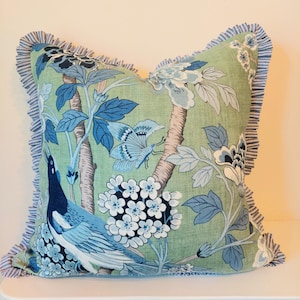 Puede incluir: Cojín decorativo cuadrado con fondo verde y estampado floral y de aves en azul y blanco. El cojín tiene un borde con volantes a rayas azules y blancas. El diseño incluye un pavo real, mariposas y flores.