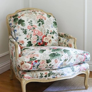May include: A vintage-style armchair with a cream-colored wooden frame. The chair is upholstered with a floral fabric featuring pink, red, and blue flowers with green foliage on a white background. The chair has a curved back and armrests.