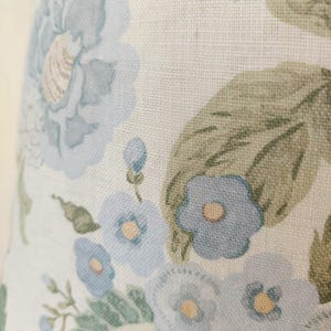 May include: Close-up of a floral pattern featuring large, light blue flowers with yellow centers and green leaves on a cream-colored background. The fabric has a textured appearance, suggesting a linen or similar material.